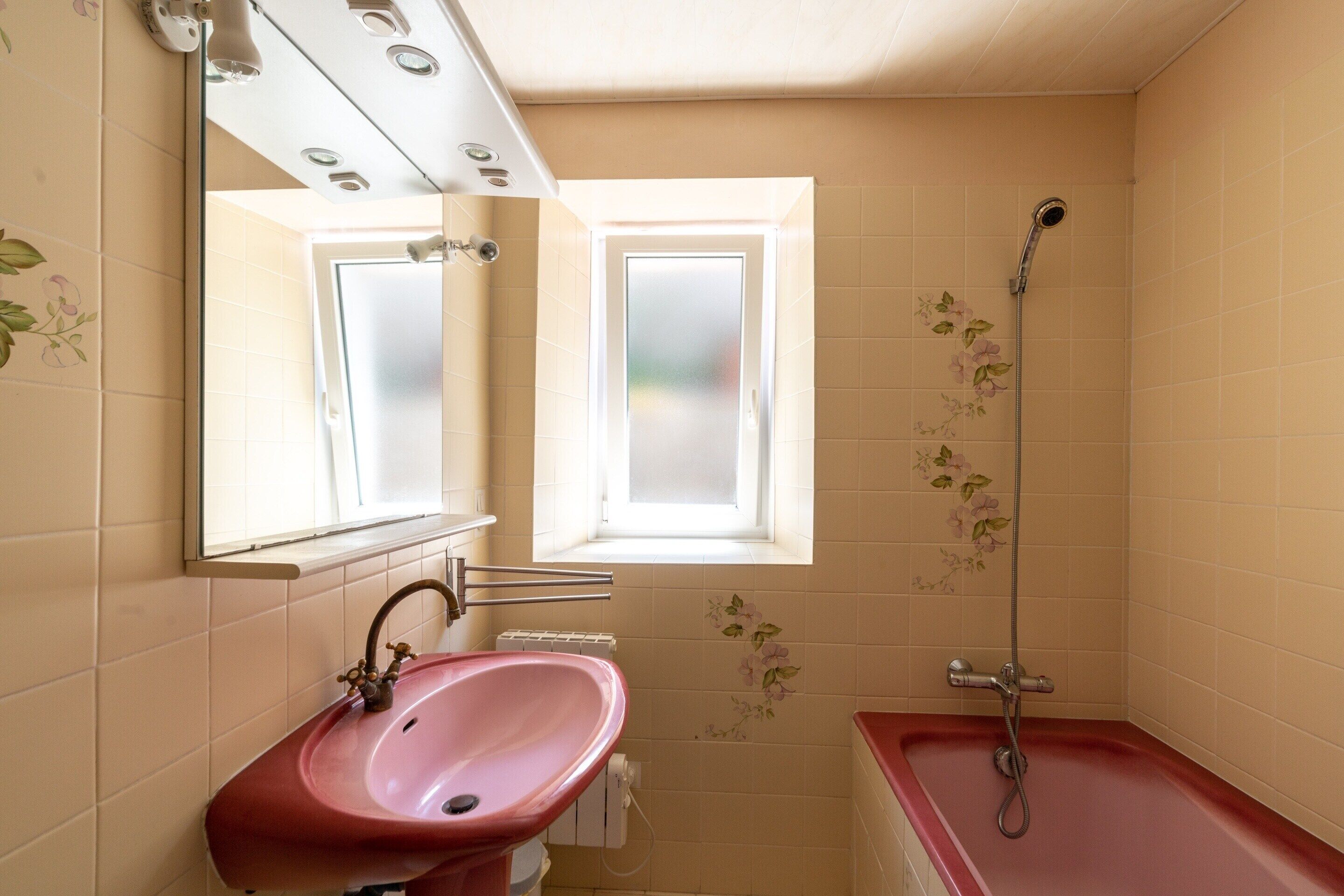 Een badkamer met een roze wastafel, een spiegel, een raam en een douche.