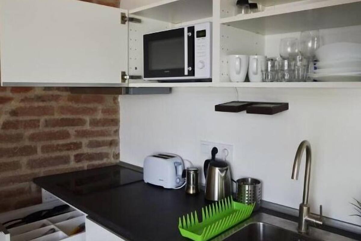 A compact kitchen with a microwave, toaster, kettle, and a green dish rack.