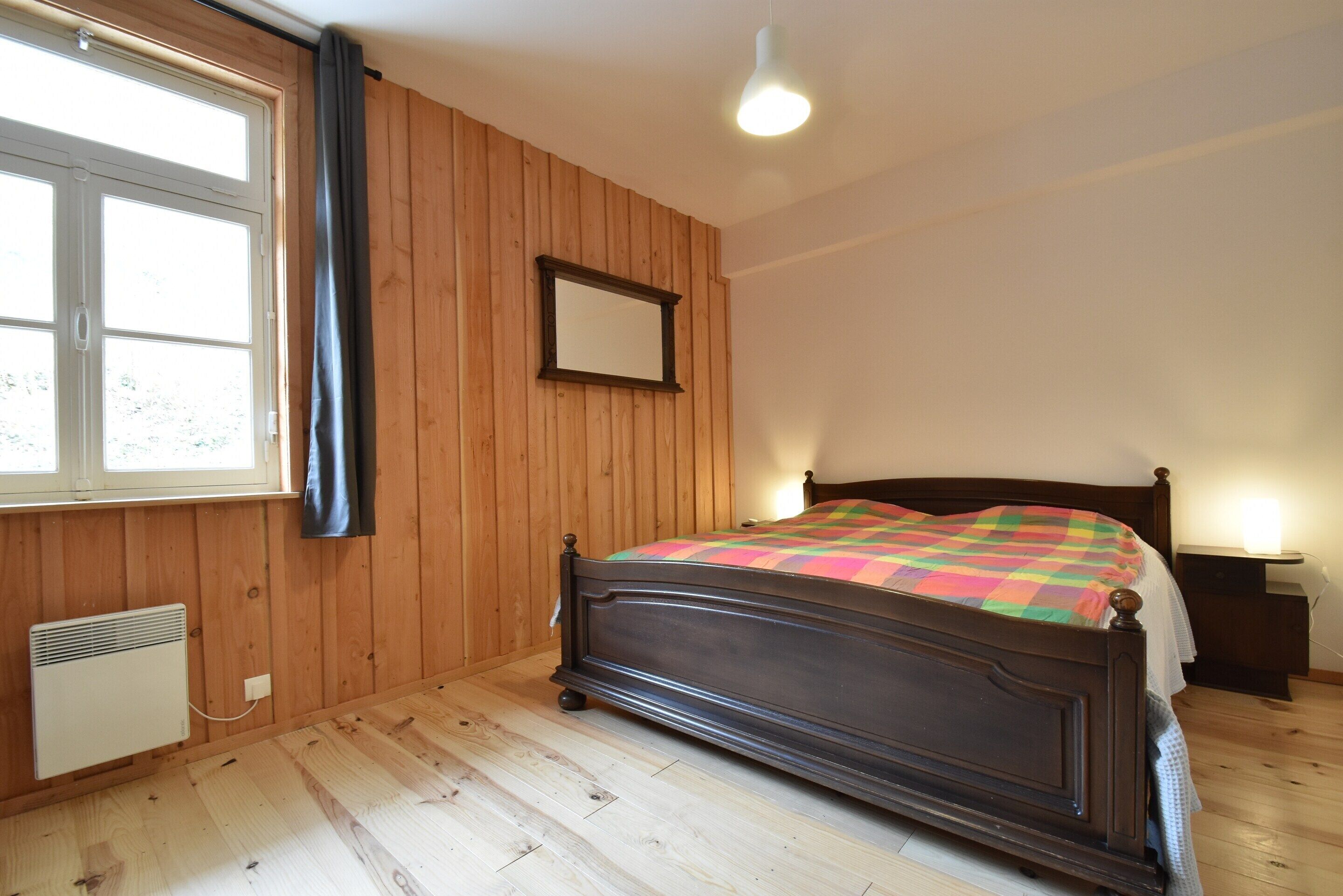 Una camera da letto con parete rivestita in legno, un letto con coperta colorata, un comodino con lampada e una finestra con tende.