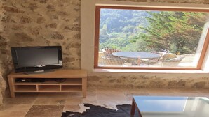 A room with a television on a wooden stand, a glass table, a cowhide rug, and a view of a patio with chairs and a table. - Villa in Ampus With Panoramic Views & Pool (Ampus)
