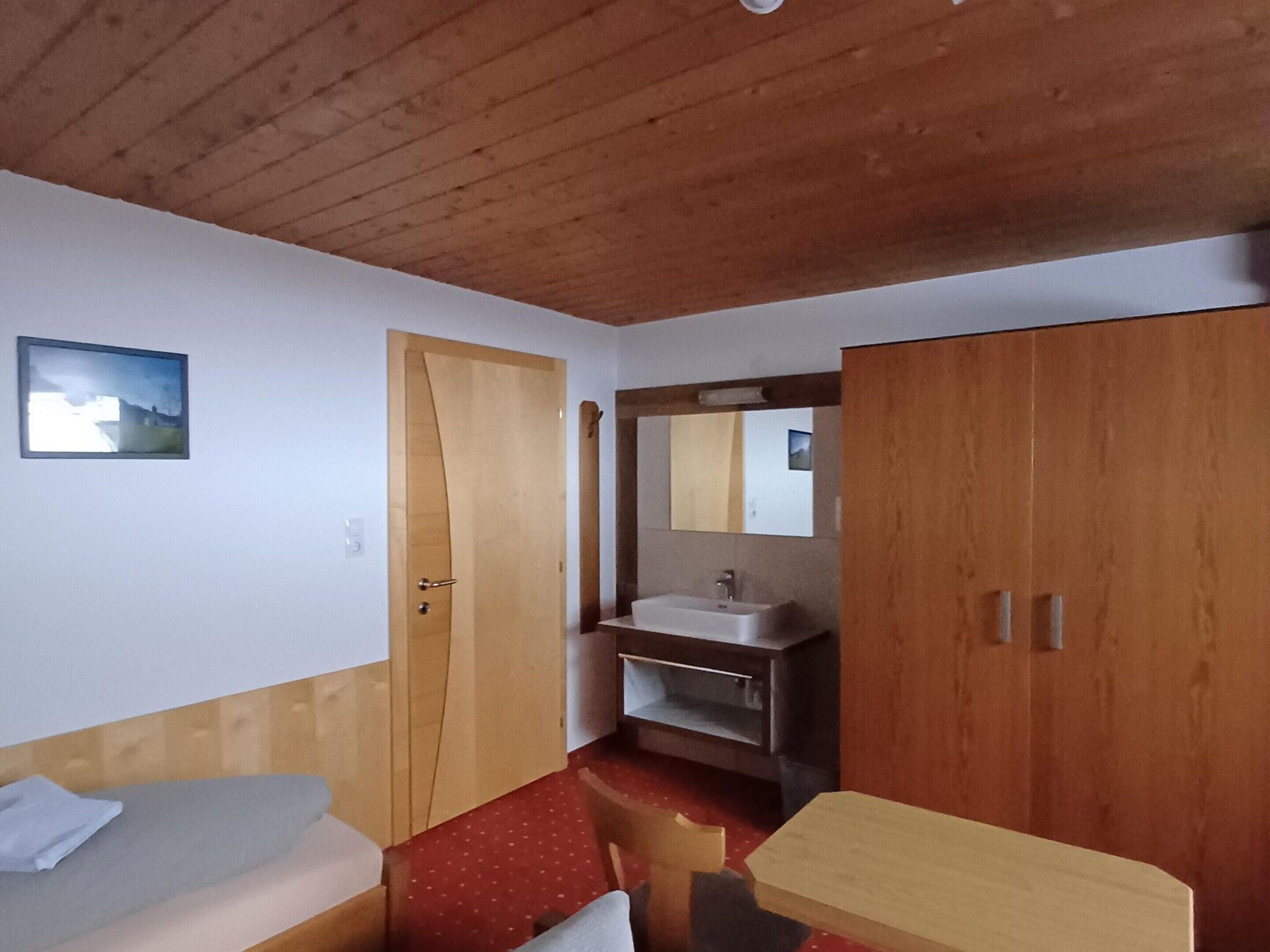 A room with a wooden ceiling, a bed with a white mattress, a red patterned carpet, a wooden wardrobe, a wooden table, a mirror, and a bathroom with a sink and mirror.