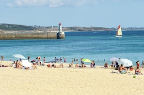 Ein Strand mit badenden Personen, einem Leuchtturm und einem Segelboot im Wasser.