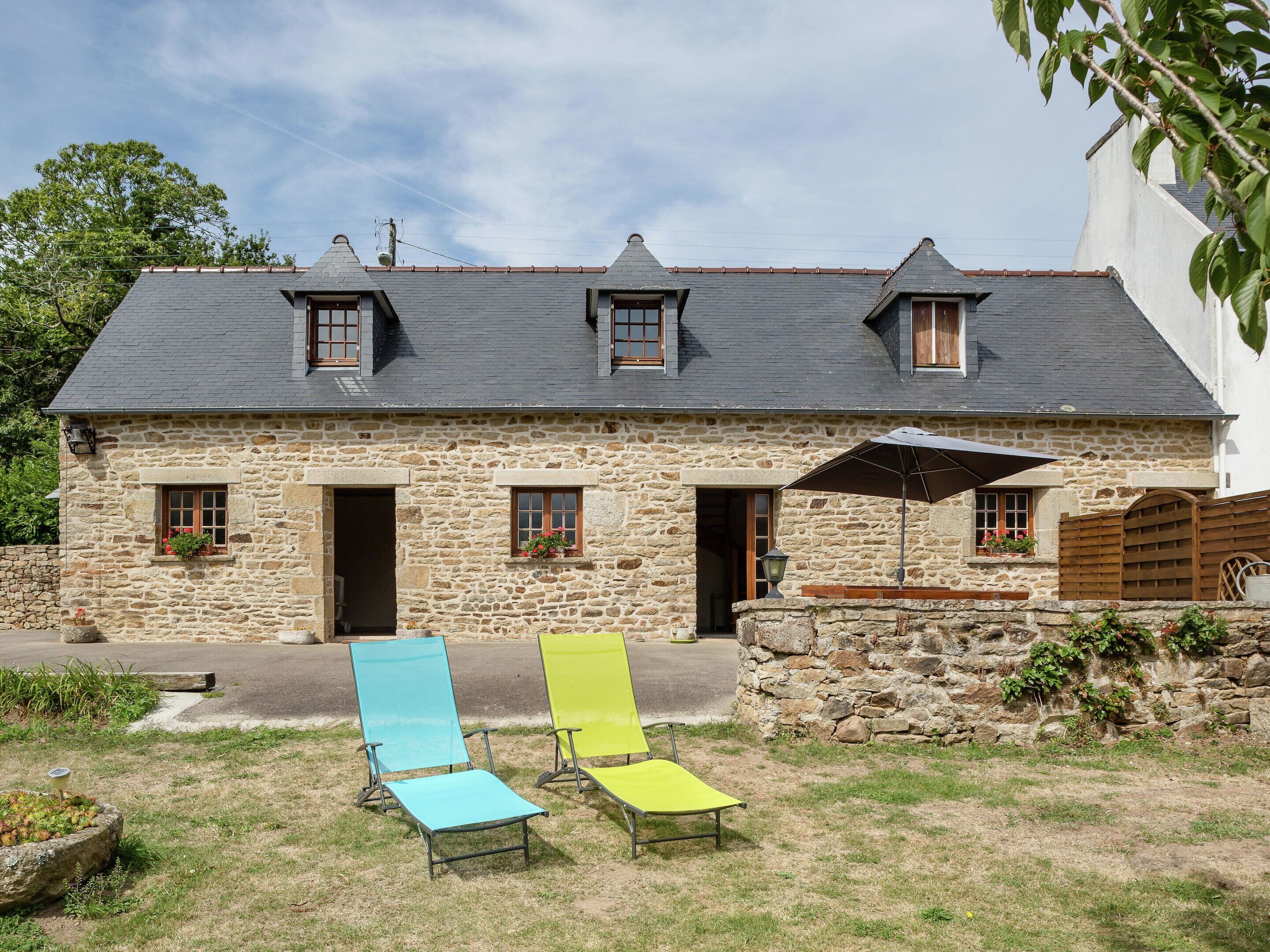 A stone building with a sloped roof, two windows, and a door. There are two lawn chairs and a patio umbrella.