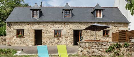 A stone building with a sloped roof, two windows, and a door. There are two lawn chairs and a patio umbrella.