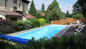 A swimming pool with a wooden deck and lounge chairs, surrounded by lush greenery and a house with a sloped roof. - Mansion in Vecoux With Pool and Garden (Vecoux)
