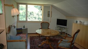 A room with a round wooden table, two wooden chairs, a sofa with cushions, a television, and a window with a view of greenery. - Charming Chalet Near Trails (Mörel-Filet)