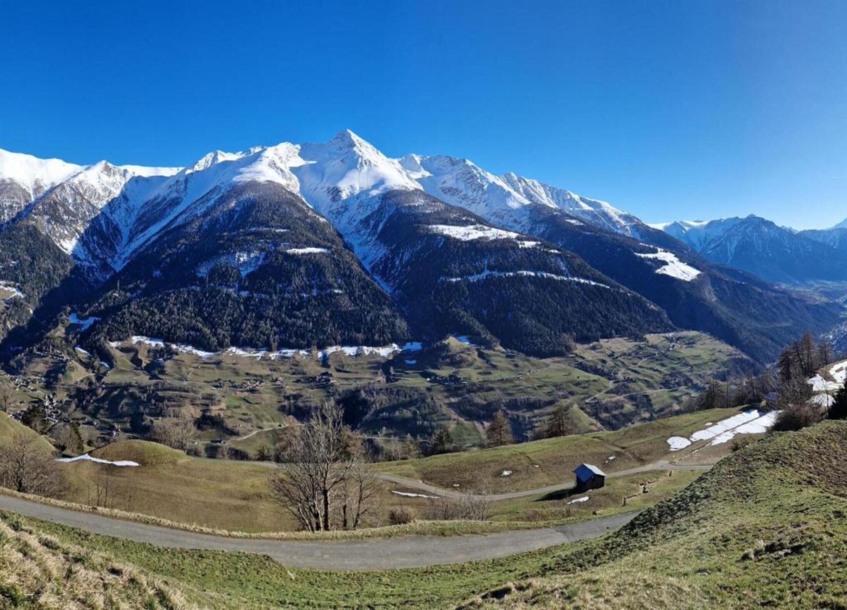 A mountainous landscape with snow-capped peaks, a winding road, and a small building in the foreground.