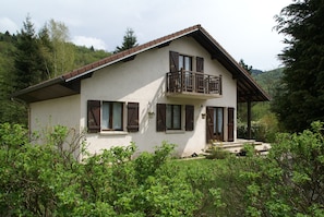 Une maison à deux étages dotée d’un balcon, entourée de verdure et de collines.
