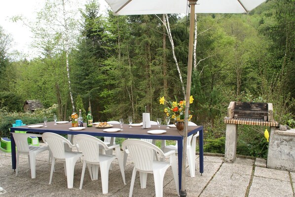 Un espace extérieur aménagé pour les repas, avec une table dressée pour six personnes, entourée de chaises en plastique blanches, d’un barbecue et d’un parasol blanc.