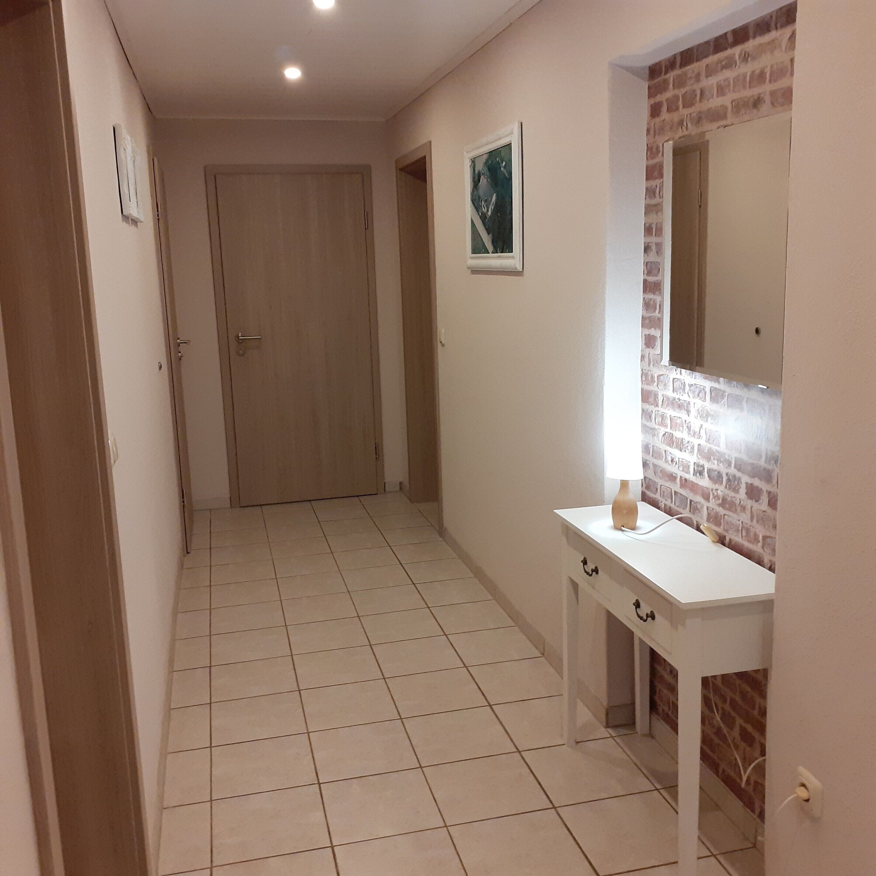 A hallway with a white tiled floor, a wooden door, a white vanity with a mirror, and a small picture on the wall.
