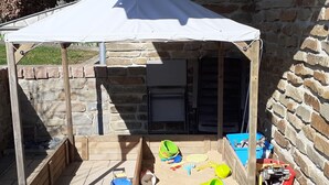 A covered outdoor play area with a stone wall backdrop, wooden play structures, and a tent providing shade.