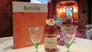 Uma garrafa de vinho Belvilla, dois copos de vinho e uma caixa de vinho Belvilla sobre uma mesa em um ambiente de madeira.