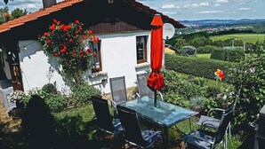 Uma casa branca com telhado vermelho, uma mesa de vidro com cadeiras e um guarda-sol vermelho.