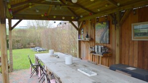 Un espace extérieur abrité, aménagé pour les repas, avec une longue table et des chaises, entouré d’une clôture en bois et d’un chemin en briques.