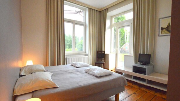 Une chambre à coucher avec un lit, un téléviseur posé sur un meuble et une fenêtre avec des rideaux.