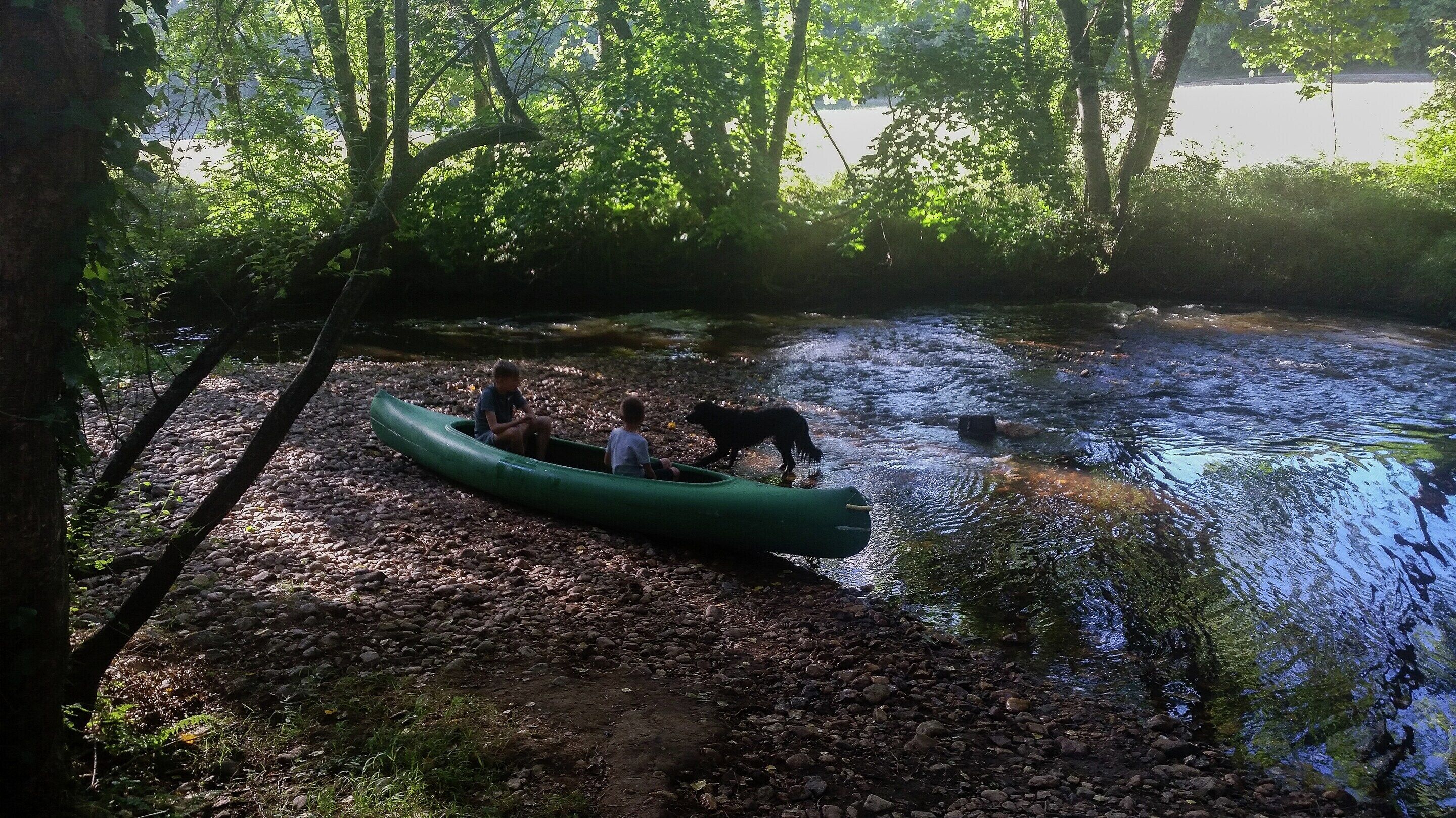 Orilla de río con un canoa verde, un perro y un niño.