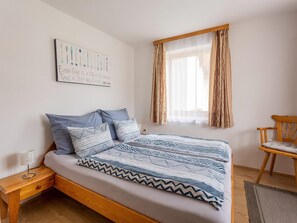 A neatly made bed with patterned bedding, a bedside table with a lamp, a wooden chair, and a window with sheer curtains.