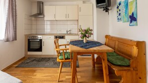 A compact living space with a kitchenette, a dining table with chairs, a wooden bench with a cushion, and a television mounted on the wall. - Apartment in Tropolach Near Nassfeld (Hermagor-Pressegger See)