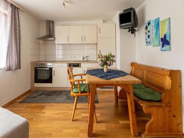 A compact living space with a kitchenette, a dining table with chairs, a wooden bench with a cushion, and a television mounted on the wall.
