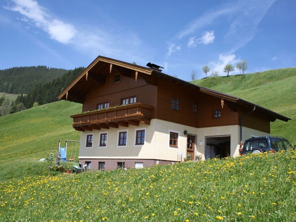 A two-story wooden house with a balcony, surrounded by a lush green landscape and a clear blue sky.