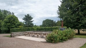 A picnic area with benches and a stone wall, surrounded by trees and a gravel path. - Holiday Home in Rogery With Sauna (Gouvy)