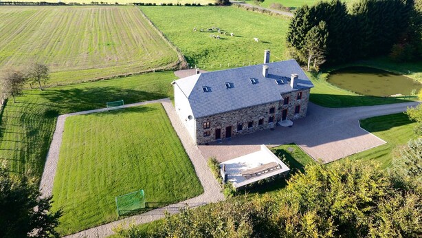 An aerial view of a stone building with a sloped roof, surrounded by green fields and a pond. - Holiday Home in Rogery With Sauna (Gouvy)