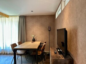 A modern dining area with a wooden table and chairs, a vase with flowers, a floor lamp, and a TV on a stand.