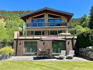 Una casa moderna de dos pisos con un balcón, ventanas amplias y un patio con mobiliario exterior.