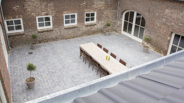 Une cour avec une longue table et des chaises, des plantes en pot et un bâtiment en briques doté de fenêtres.