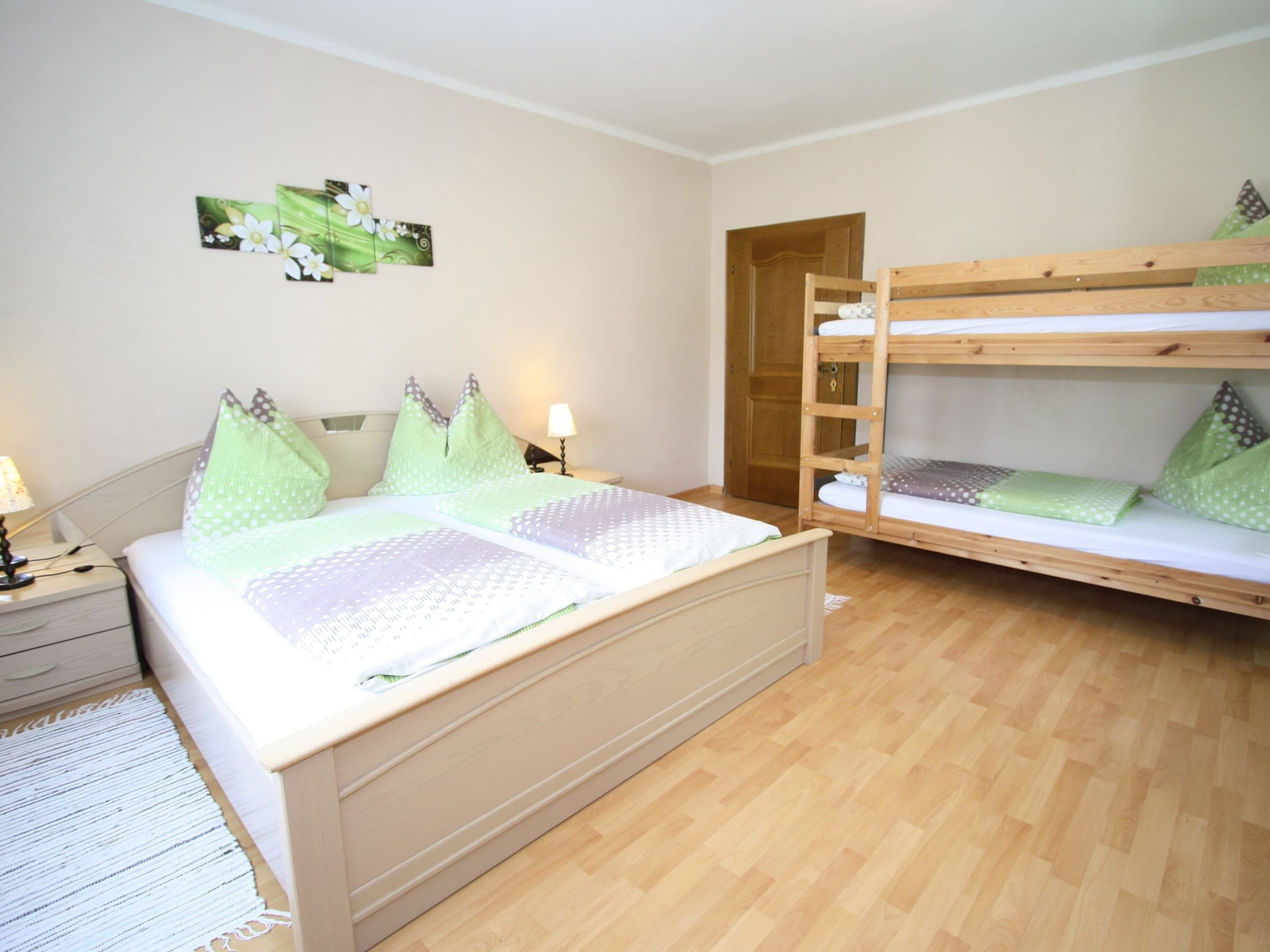A bedroom with a wooden bunk bed, a single bed with patterned bedding, a nightstand with a lamp, and a wall-mounted artwork.