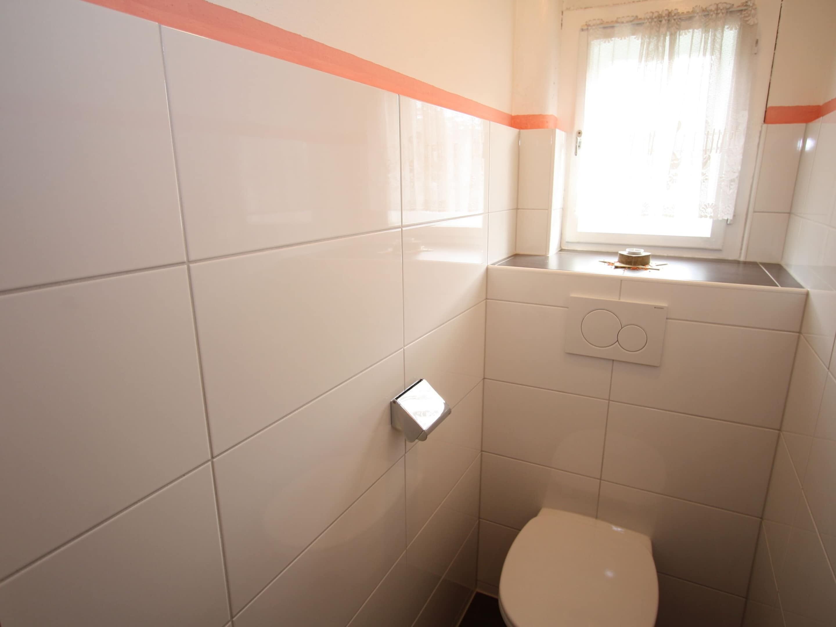 A small, clean bathroom with white tiles, a toilet, and a window with a curtain.