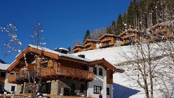 A chalet-style building with a wooden balcony, surrounded by snow-covered trees and a clear blue sky.