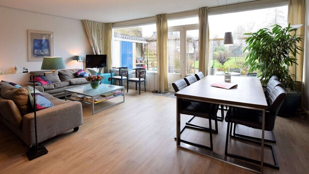Una sala moderna con sofá, mesa de centro y mesa de comedor. Hay un televisor, una lámpara y una planta. La habitación tiene ventanas amplias con cortinas.