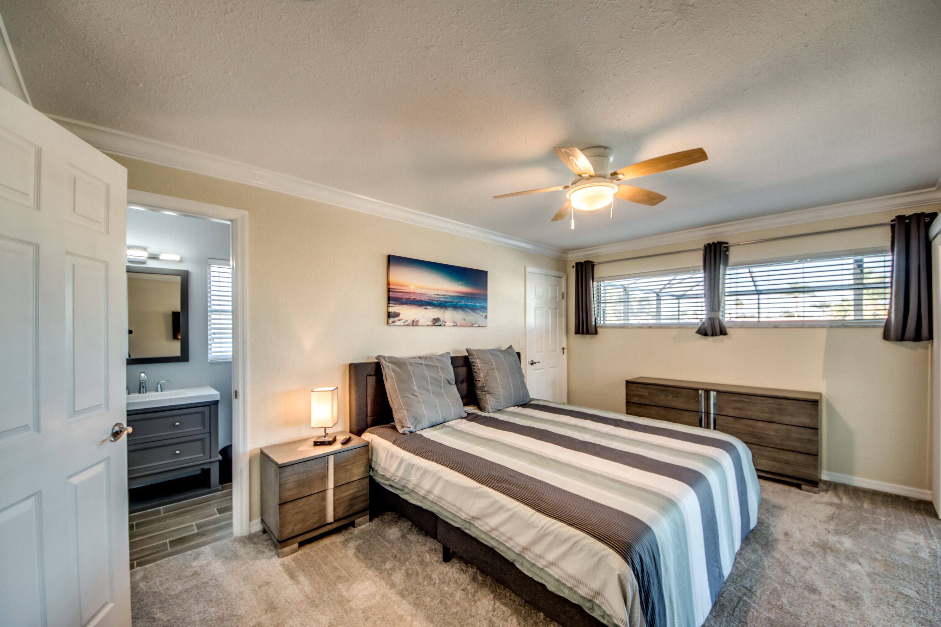 A bedroom with a bed, a nightstand, a ceiling fan, a window with curtains, and a bathroom with a mirror and vanity.