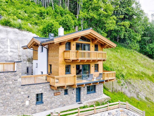 Eine mehrstöckige Holzhütte mit Balkonen, Steinmauern und Blick auf einen grünen Hang.