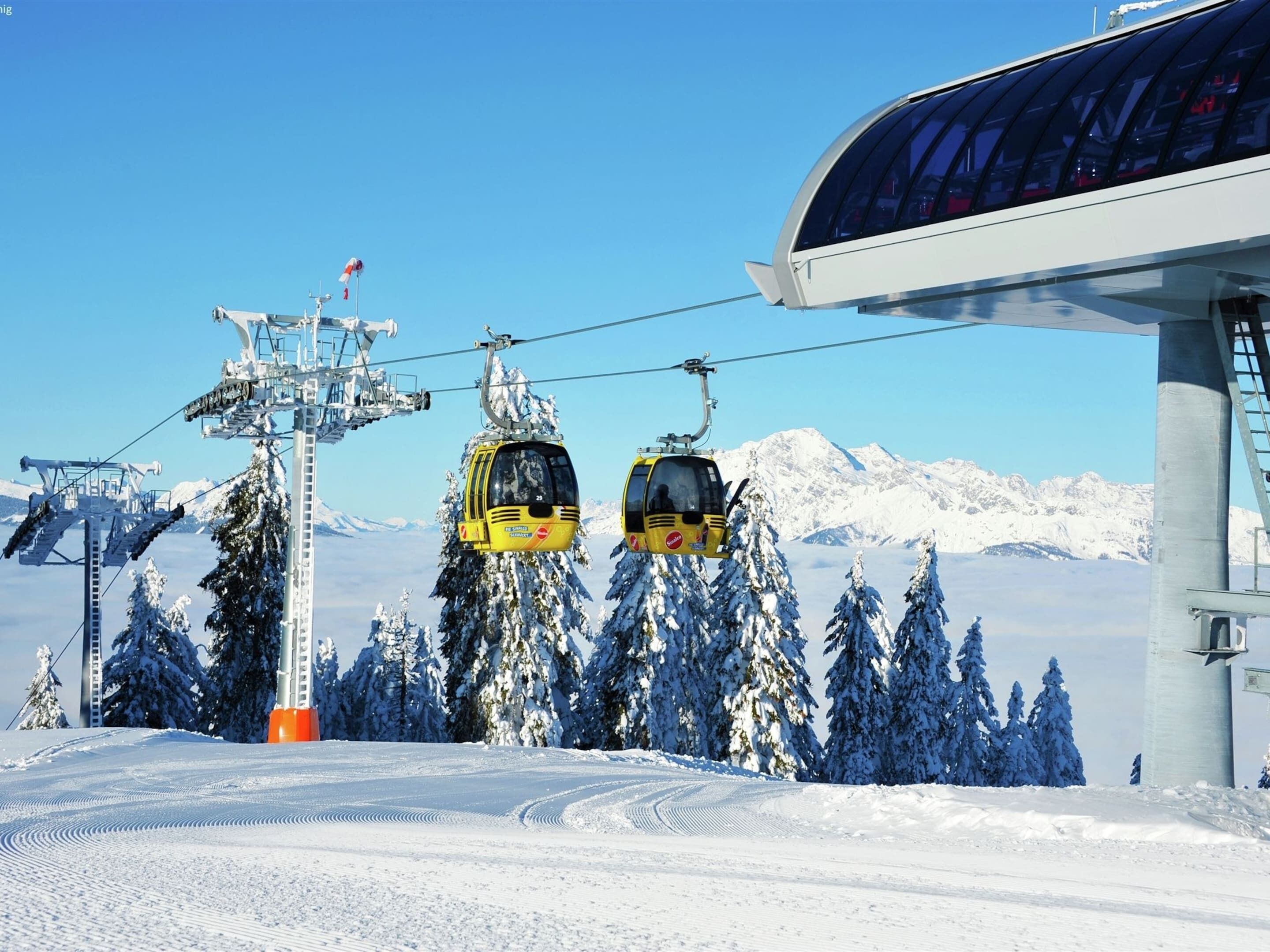 A ski lift with two yellow cabins, a cable system, and snow-covered trees against a mountain backdrop.