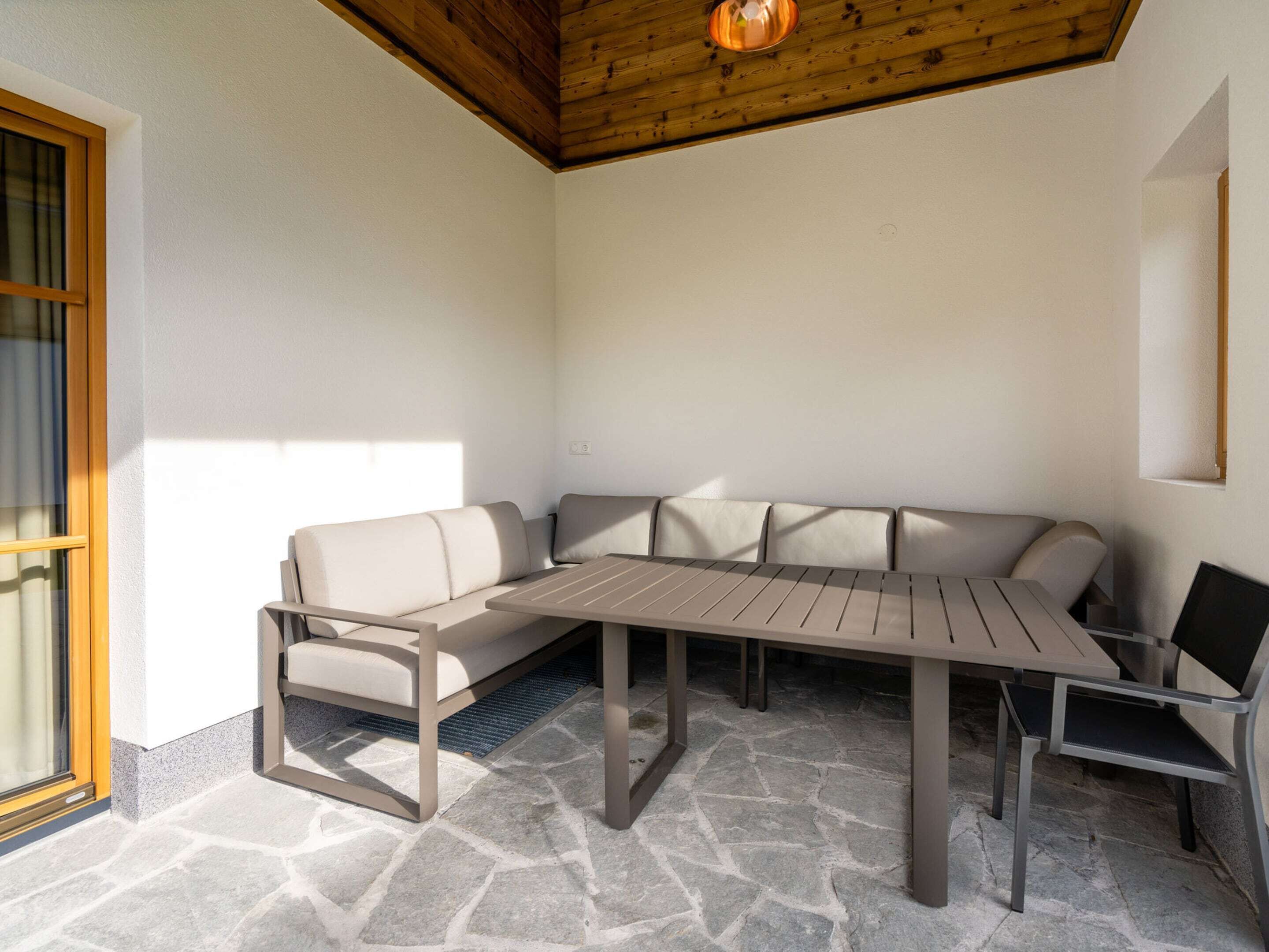Een zonnig terras met een stenen vloer, houten plafond en een beige bank en tafel.
