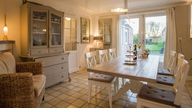 Een eetruimte met een houten tafel en witte stoelen, een kast met glazen deuren en uitzicht op de tuin door grote ramen.