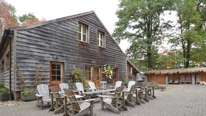 A rustic wooden building with a thatched-roof structure, outdoor seating with Adirondack chairs, and a paved courtyard. - Lodge Maasduinen Near National Park (Wellerlooi)