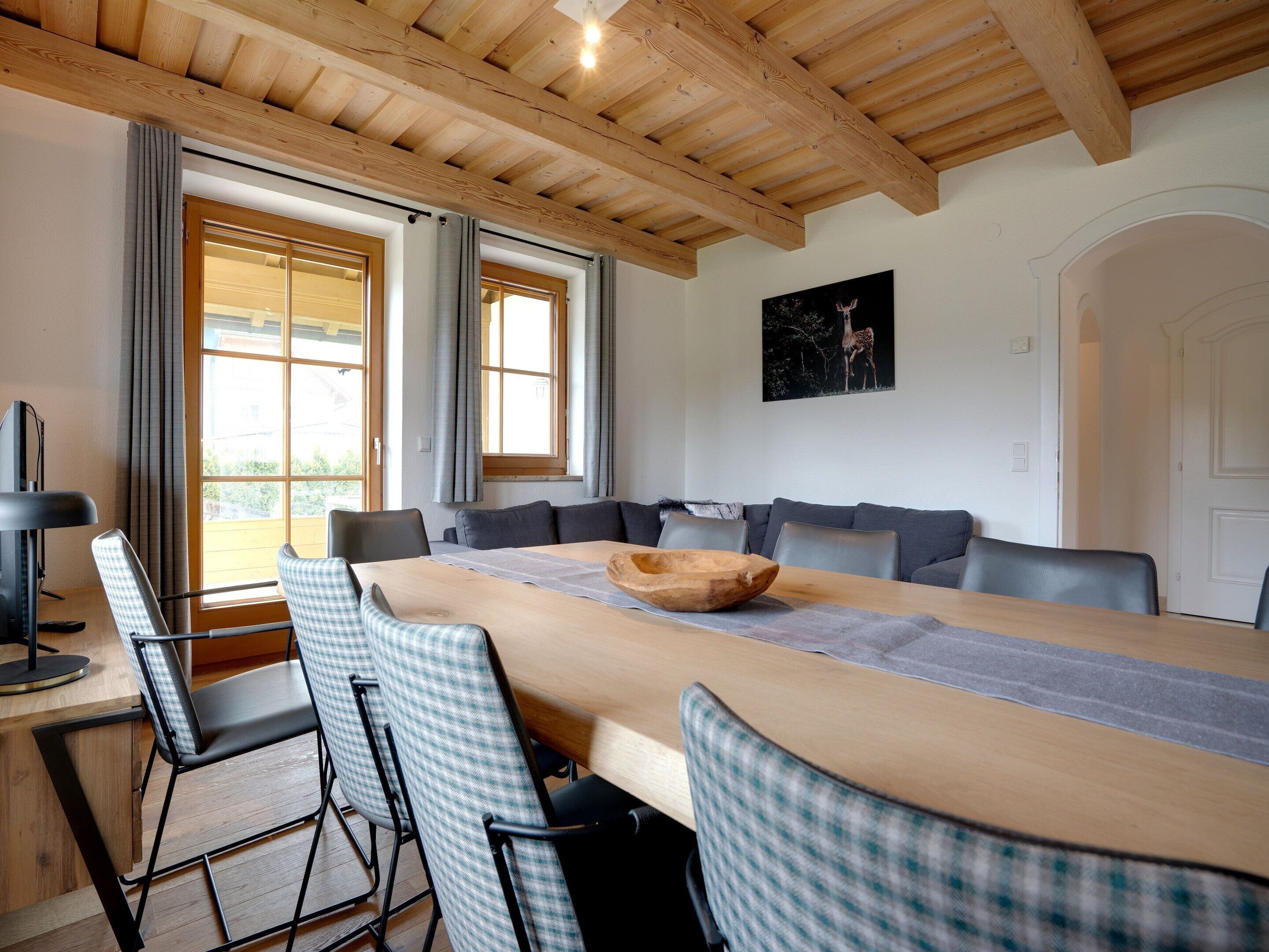 Een eetgedeelte met een lange tafel, geruit gestoffeerde stoelen, een houten plafond en een groot raam met gordijnen.