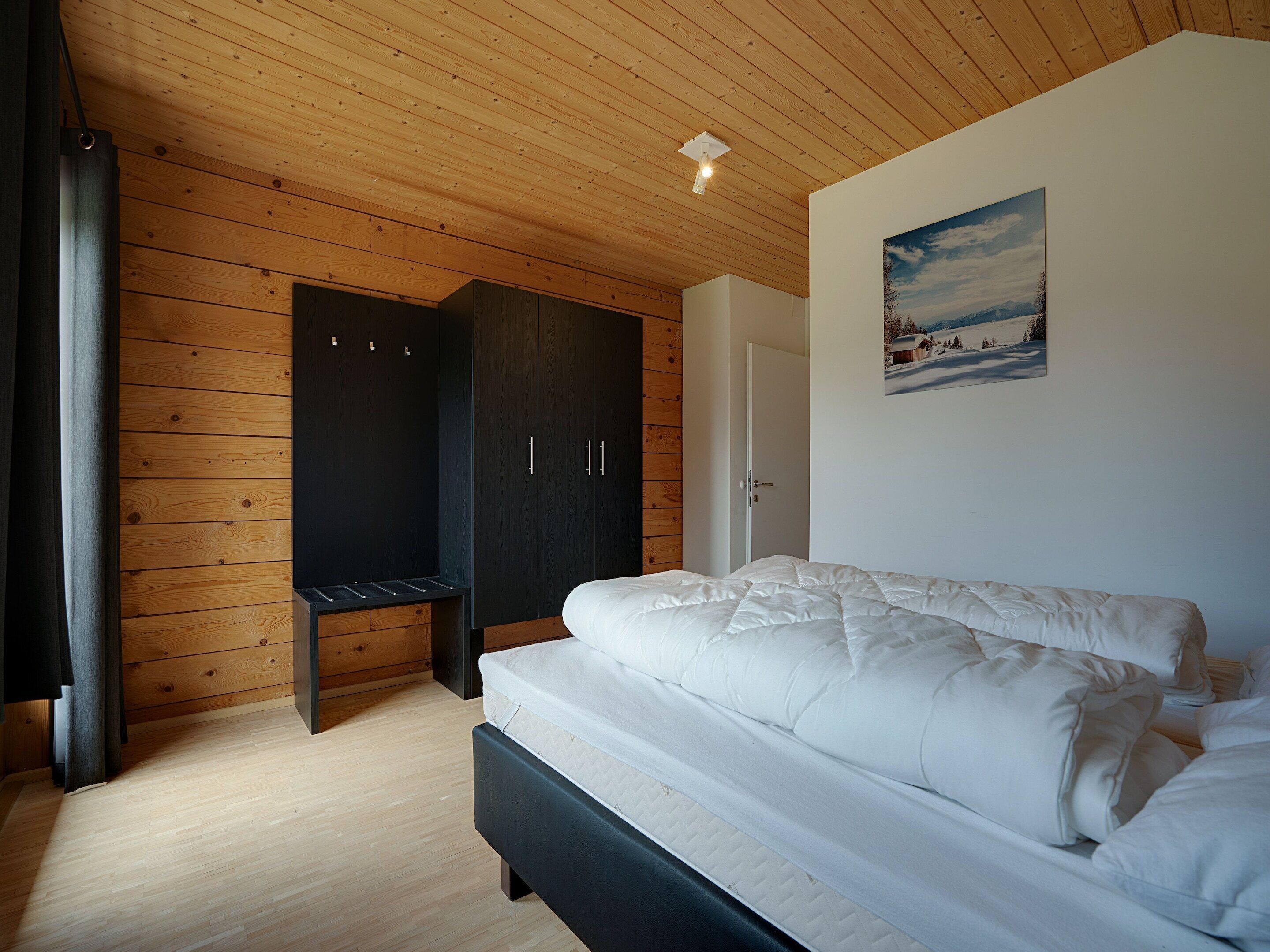 Quarto com teto de madeira, cama com roupa de cama branca, guarda-roupa e porta que dá para outro cômodo.