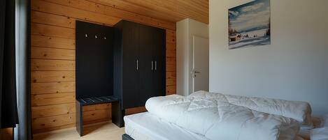Quarto com teto de madeira, cama com roupa de cama branca, guarda-roupa e porta que dá para outro cômodo.