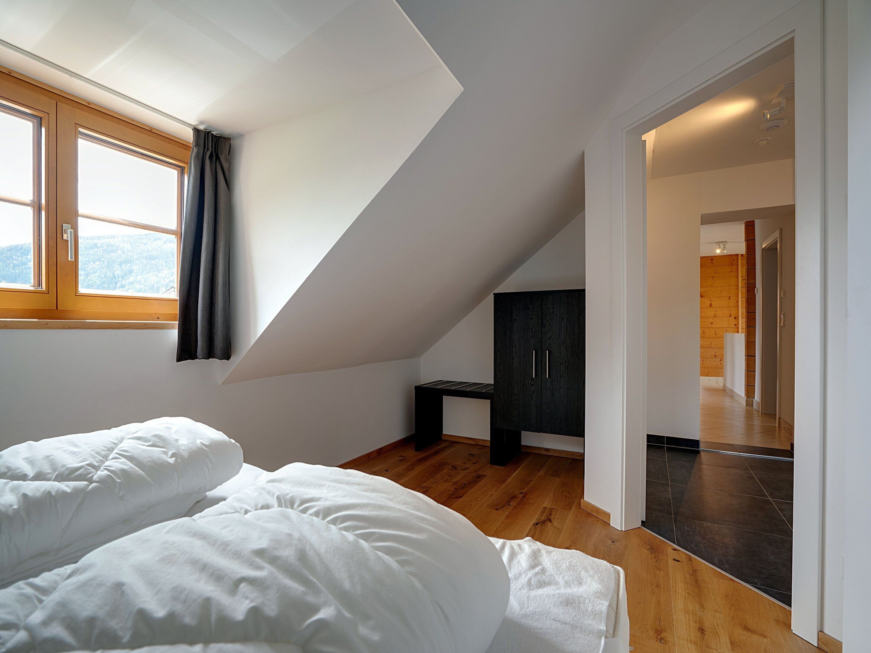 A bedroom with a sloped ceiling, a bed with white bedding, a dark wooden wardrobe, and a view through a window.