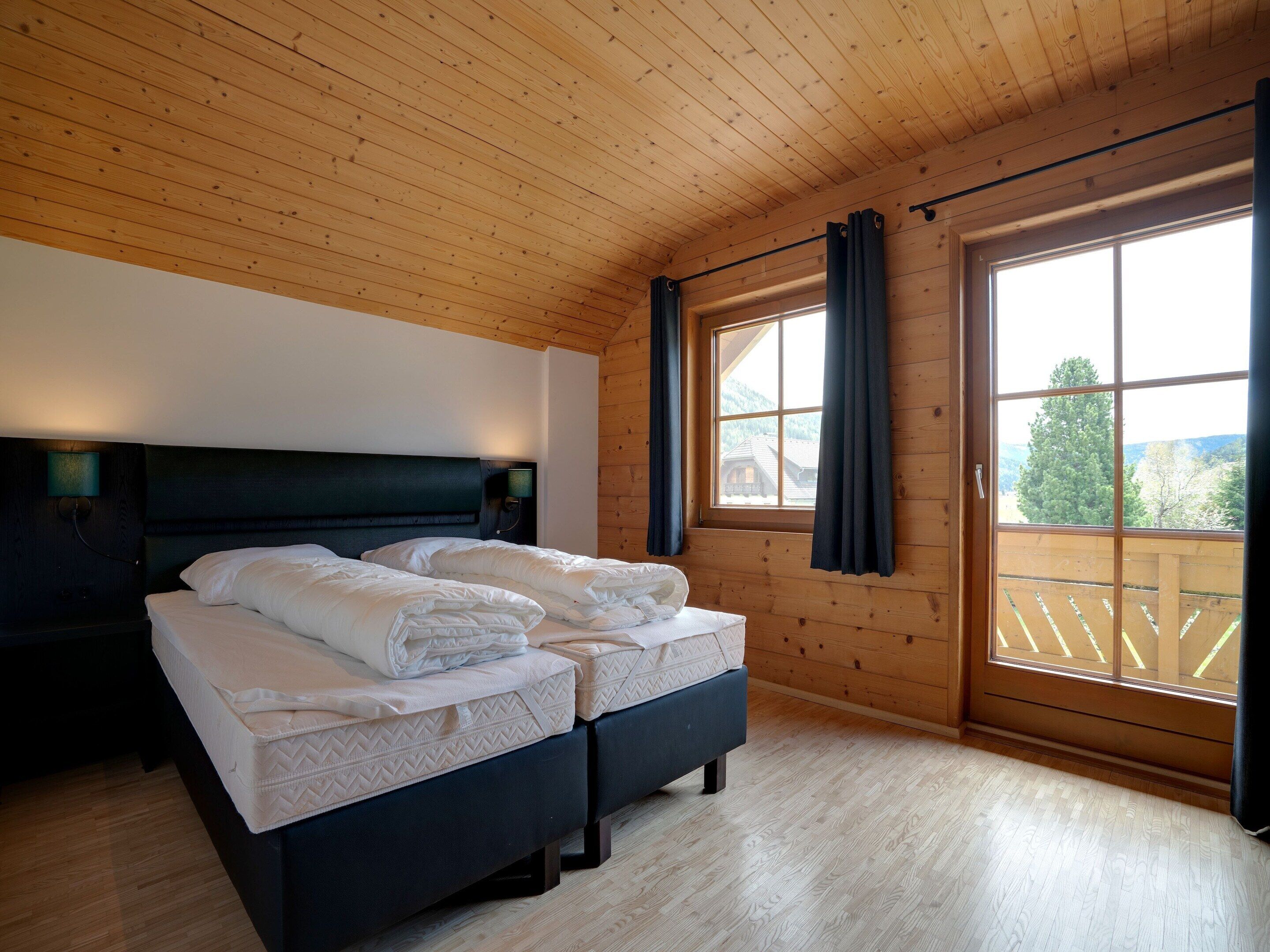 A bedroom with two beds, wooden ceiling, large window with curtains, and a view of the outside.