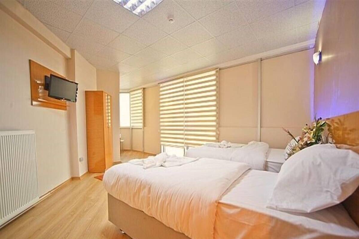 A modern bedroom with a large bed, a TV mounted on the wall, and a window with blinds.