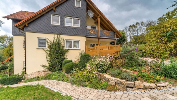 A two-story house with a sloped roof, a wooden deck, and a stone pathway leading to the entrance. - Apartment in Marktrodach With Sauna & Garden (Marktrodach)