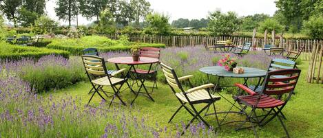 An outdoor garden with a round table and chairs, surrounded by lush greenery and purple flowers.