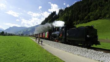 A steam locomotive with passengers on a train track, surrounded by green fields and mountains.
