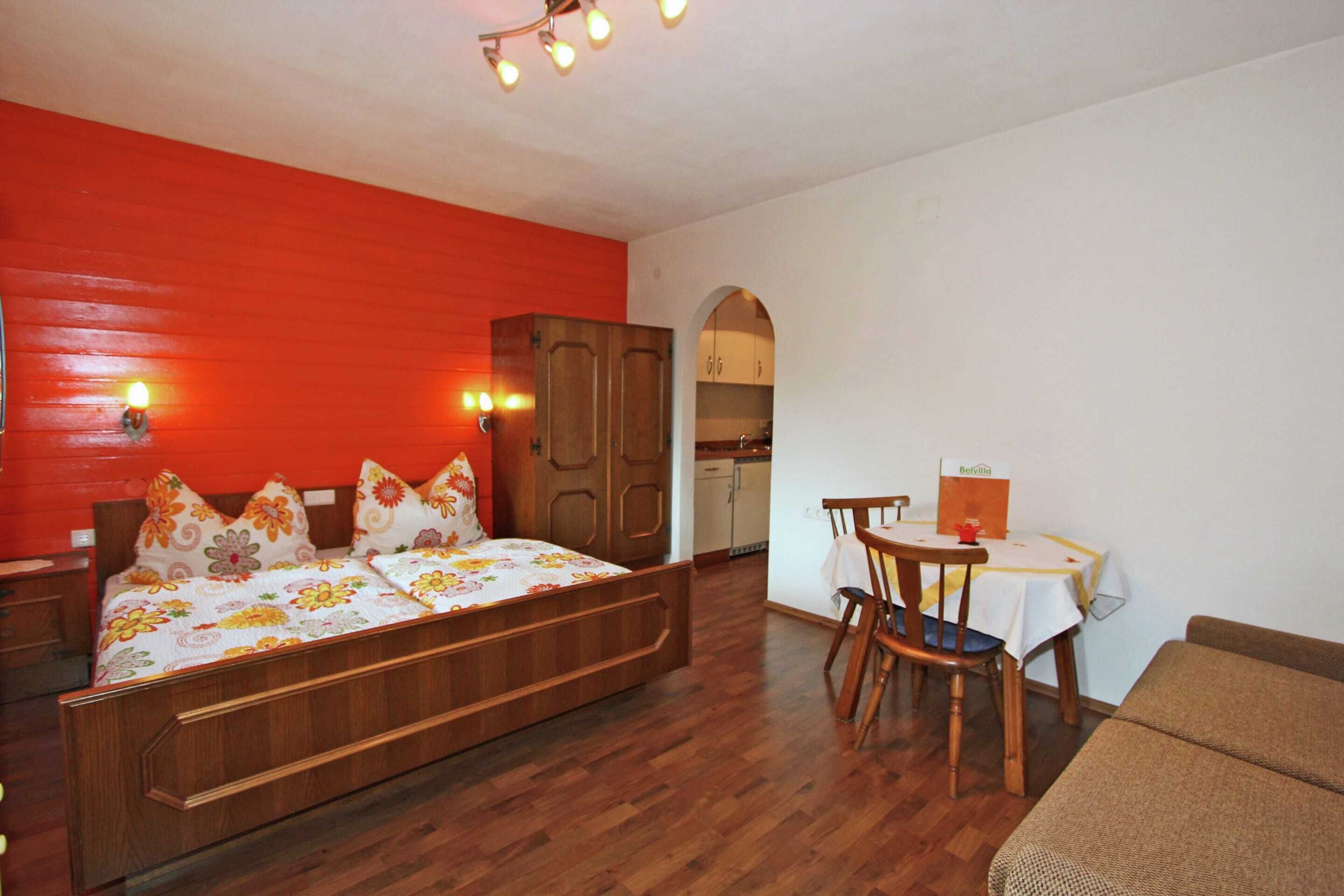 A bedroom with a wooden bed, a red accent wall, a dining area with a table and chairs, and a kitchen in the background.