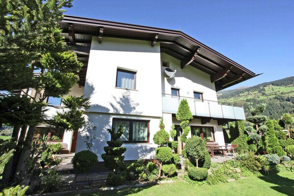 A modern two-story house with a balcony, surrounded by well-maintained gardens and trees.